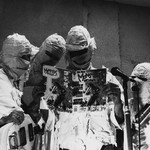 Black and white portrait of four members of The Mummies, dressed in mummy attire and collectively reading a magazine on-stage.