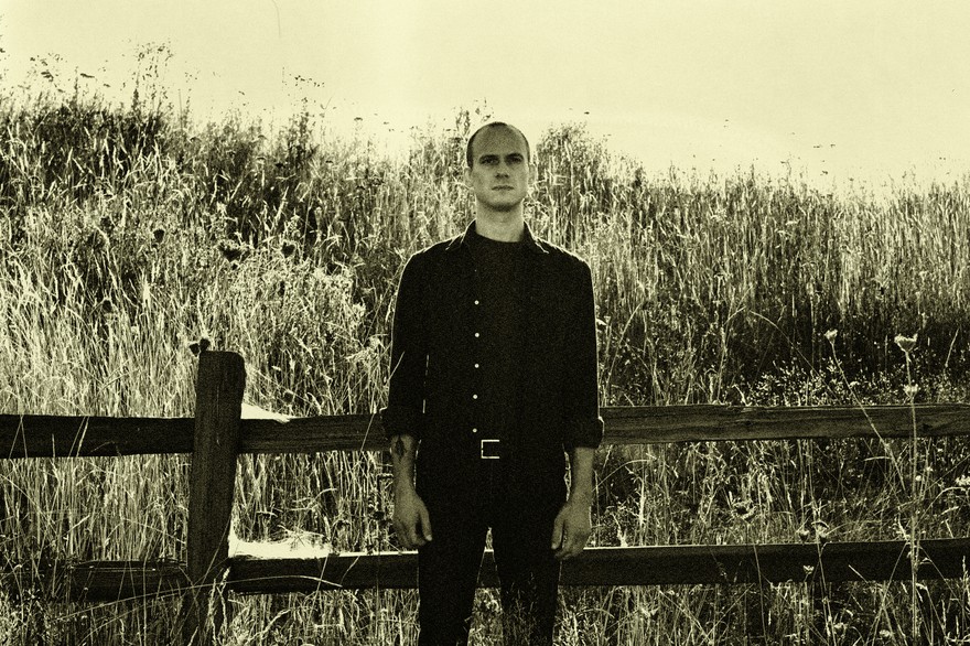 Outdoor portrait of solo artist Mizmor standing in front of a wooden fence in a prairie setting.