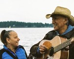 Outdoor portrait of duo Willie Thrasher & Linda Saddleback gazing toward one another, with a large body of water behind them. Willie Thrasher is holding a guitar.