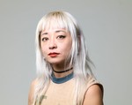 Artist Evicshen is shown posing for a colour photo in front of an off-white background. They are shown from the shoulders up, wearing a tan shirt and having bleach blonde shoulder length hair