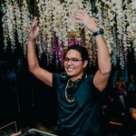 DJ BLKFT is seen performing with headphone around their neck, raising their hands in the air enthusiastically. White flowers hang behind them. 