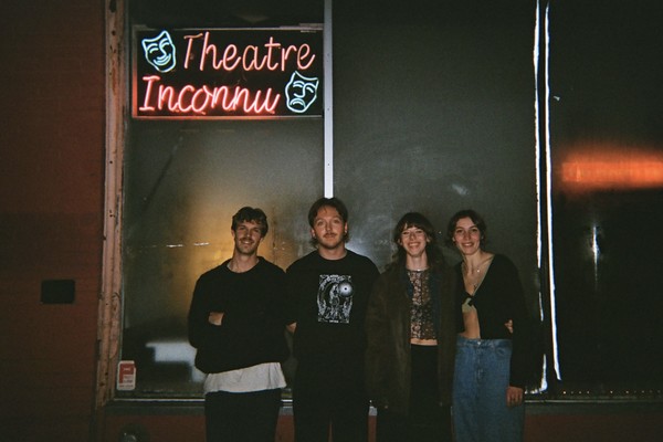 Photo taken at night, featuring four members of Blush. standing outside a window with a neon sign above which reads "Theatre Inconnu."