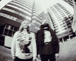 A blurry black and white photo shows two members of Genizah standing outside with two tall buildings behind them.