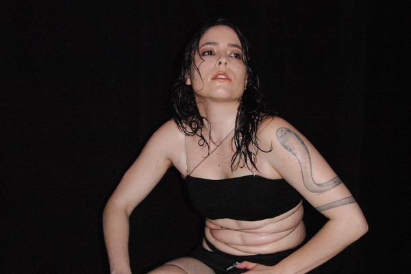 A colour photo shows artist Kee Avil crouching on a concrete surface wearing a black outfit and tight plastic strings around their stomach and legs.