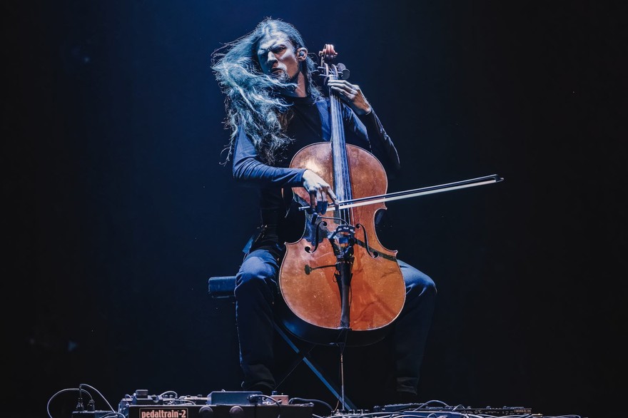 A colour photo shows cello Raphael Weinroth-Browne furiously playing a cello against a black backdrop. They are dressed all in black with long dark hair and various pedals and cords sit on the floor in front.