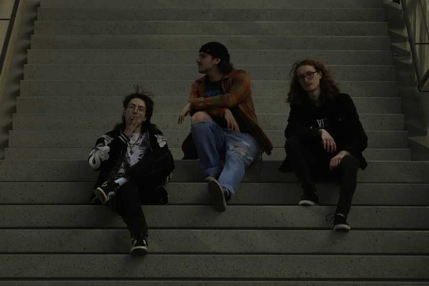 A dark lit colour photo shows three-piece group schoolyard sitting on a set of concrete stairs. The member on the left is seen smoking a cigarette, the member in the middle looks to their right, and the person on the right is looking straight at the camera.