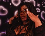 Hip-hop artist Tea Fannie poses with her hands touching her glasses in front of a bright wall with neon pink hearts