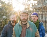 A colour photo shows the three members of UTILS standing outdoors in a lightly wooded area with leafless branches and softly lit by the setting sun in the background. The member in the foreground wears glass, a baseball cap, and a green and tan jacket. Behind them are the two other members, one wearing a dark jacket and the other wearing a blue puffer jacket, purple toque, and a green scarf.